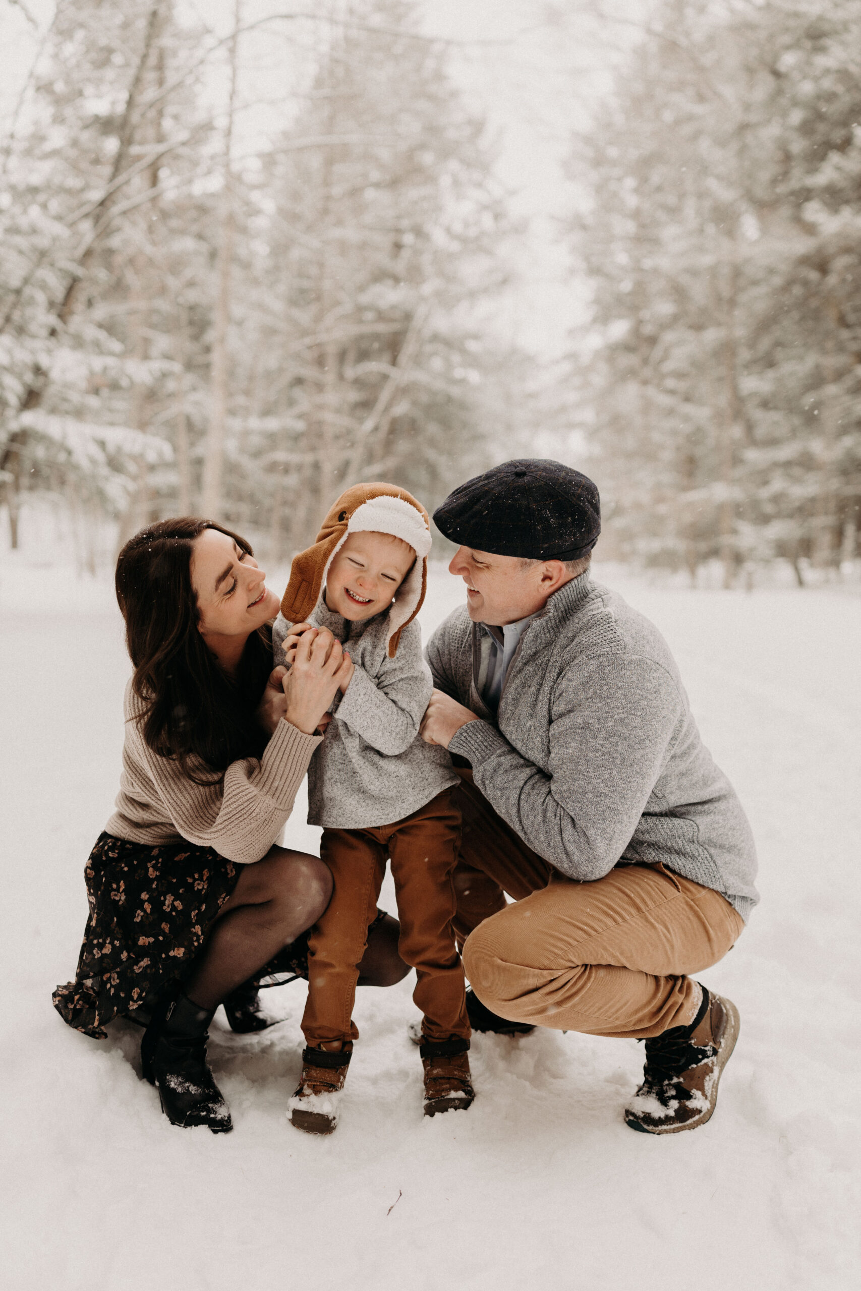 Parents kneel in the snow laughing with their young child during a winter family photo session in Vermont, surrounded by snow-covered trees.